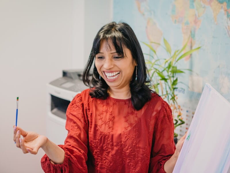 A teacher of a painting class smiles with a paintbrush and canvas in her hand.
